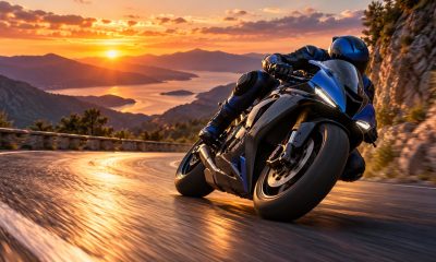 A motorcycle rider leans into a mountain-road bend above a lake at sunset.